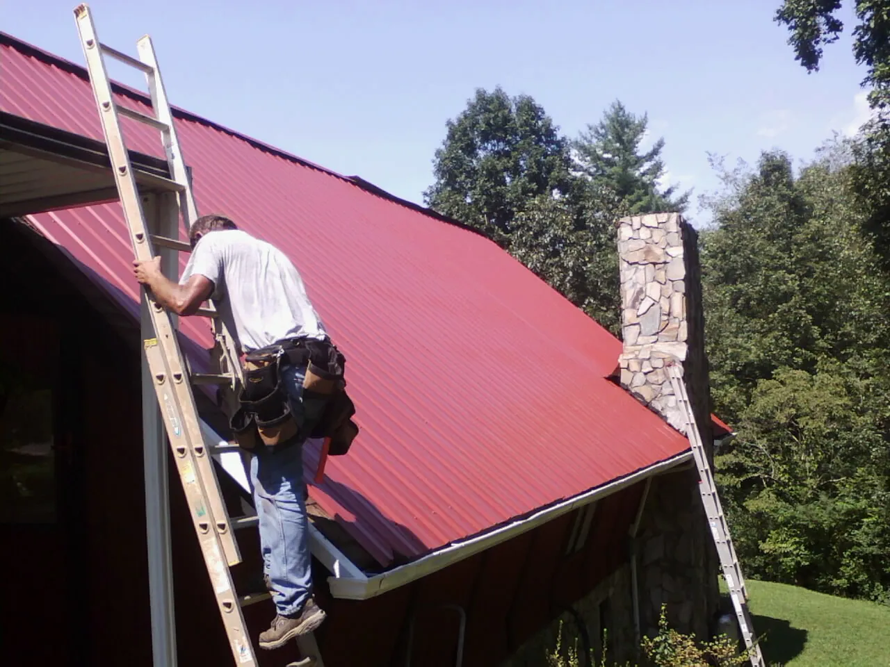 Roofer on a ladder beside a red metal roof and stone chimney — Soffit & Fascia Repair by Roofing Brothers in West Farmington